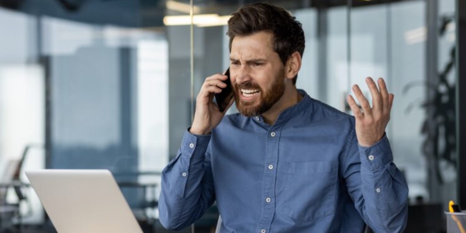 Angry and irritated young businessman sitting in the office at the desk and talking on the phone while waving his hands.