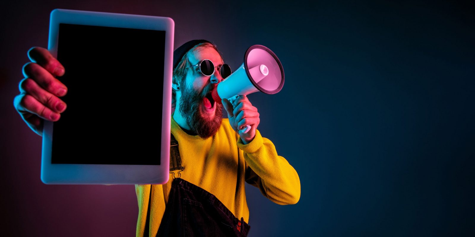 Showing tablet's blank screen. Caucasian man's portrait on gradient studio background in neon light. Beautiful male model with hipster style. Concept of human emotions, facial expression, sales, ad.